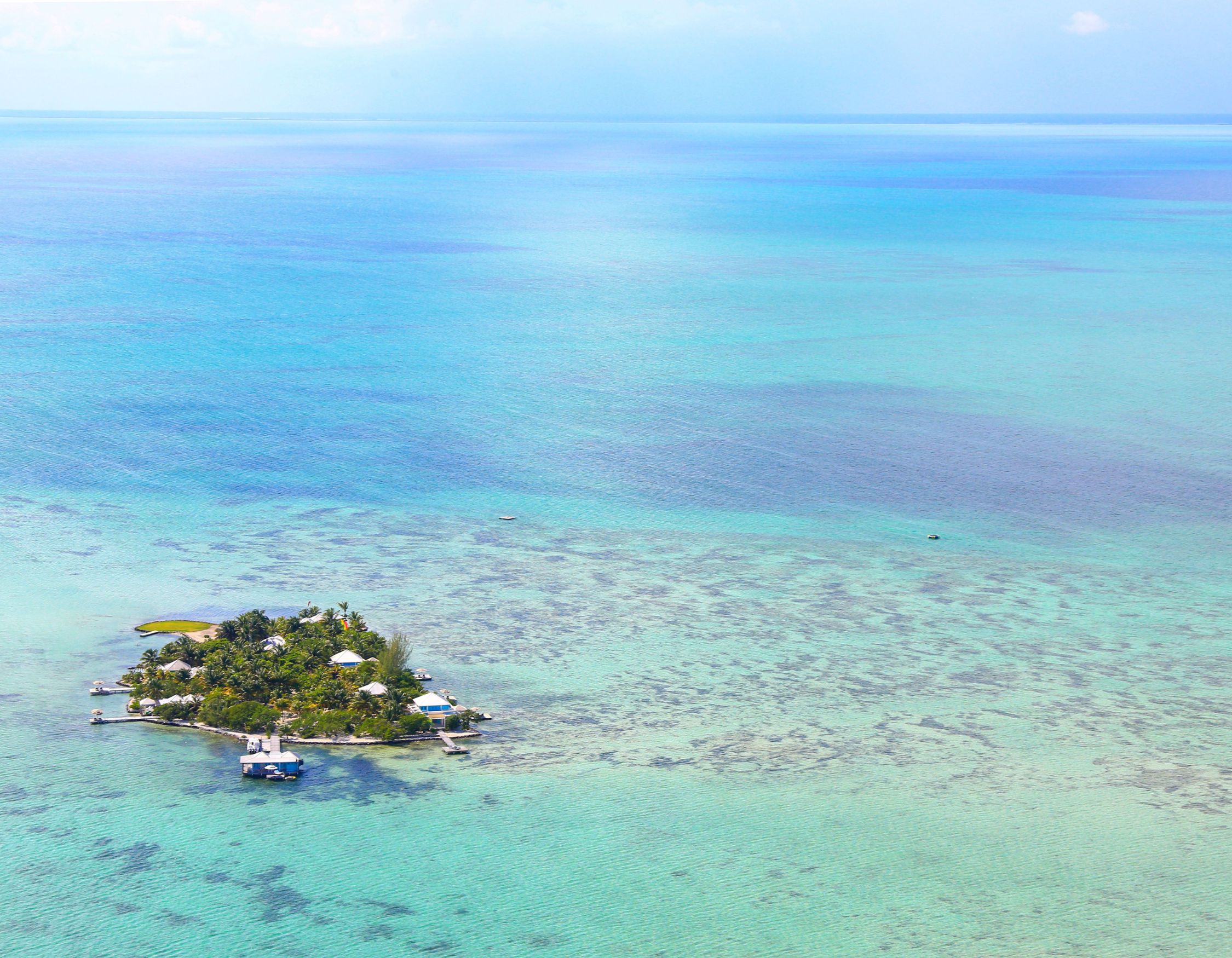 Aerial view of a small tropical island surrounded by vibrant turquoise waters and patches of coral reefs. The island features lush greenery and a few structures, with a clear blue sky overhead.