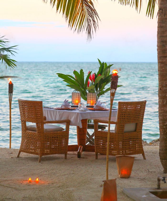 A charming outdoor dining setup by the ocean, featuring a table elegantly set for two with tropical flowers and soft lighting. Palm trees frame the scene, and tiki torches are positioned nearby, creating a romantic atmosphere against the backdrop of the water and sunset.
