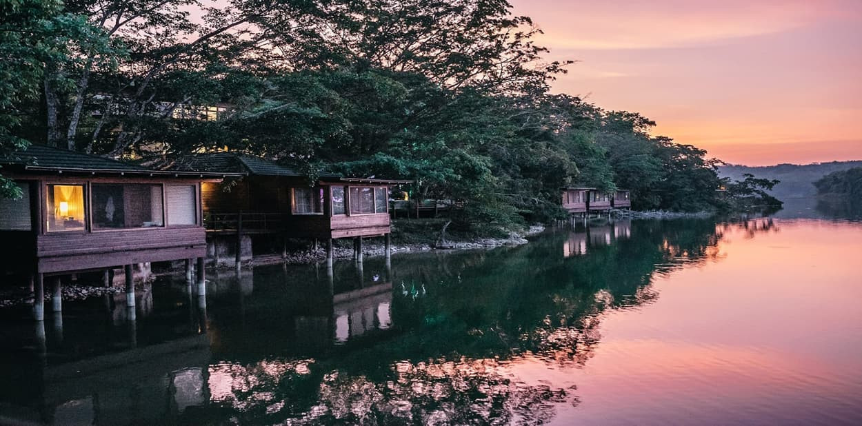 Waterside cabins reflecting on calm water at sunset, surrounded by lush trees and a serene landscape.