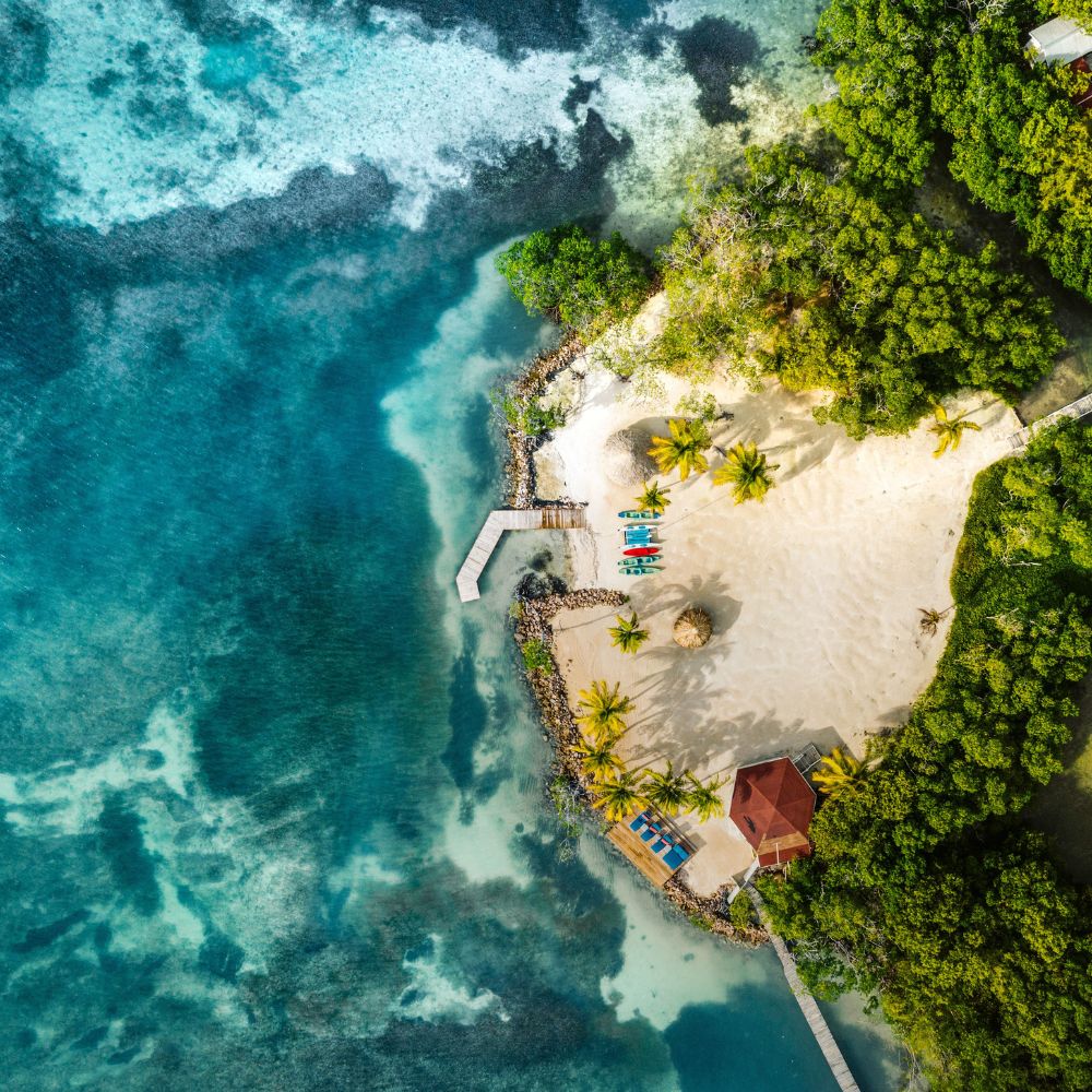A serene tropical island featuring a sandy beach, palm trees, and a small red-roofed building. Clear turquoise waters surround the island, with a wooden dock extending into the sea. Colorful kayaks are lined up on the shore.