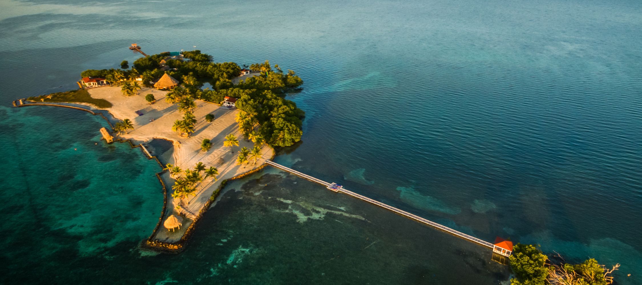 Aerial view of a tropical island with sandy beaches, palm trees, and a wooden dock extending into the turquoise water. A thatched-roof structure is visible on the island, surrounded by a calm sea.