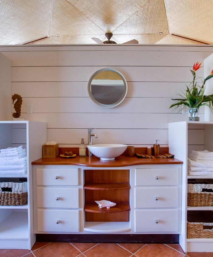 A bright and airy bathroom featuring a wooden countertop with a round sink, flanked by white cabinetry and open shelving. Neatly stacked towels and decorative baskets are arranged on either side. A circular mirror reflects the ceiling's design, and a potted plant adds a touch of greenery.