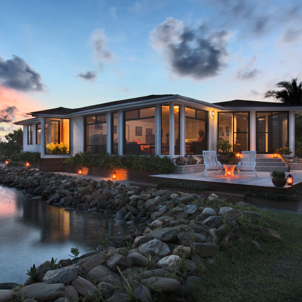 A modern house with large glass windows sits near a calm body of water, surrounded by lush greenery and rocks. Soft lights illuminate the outdoor area, creating a warm ambiance at twilight.