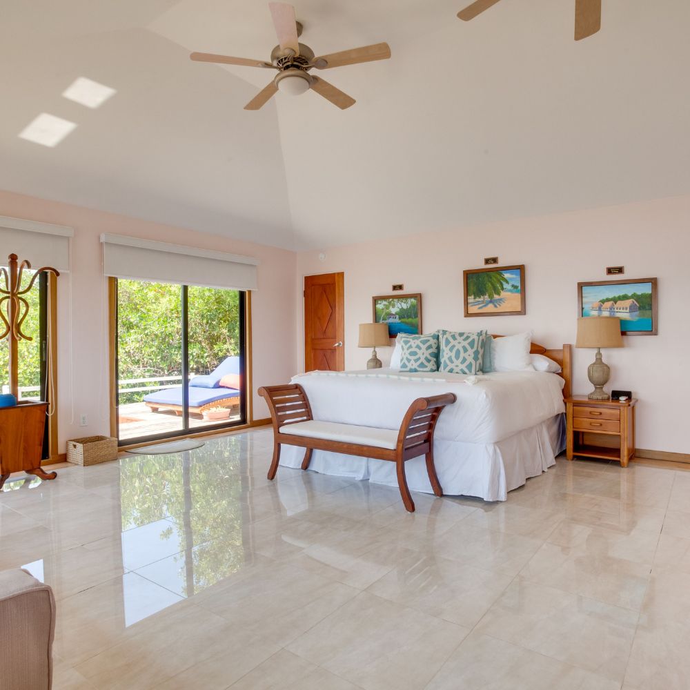 Bright and airy bedroom featuring a large bed with decorative pillows, a wooden bench at the foot, and two paintings on the wall. A sliding glass door leads to an outdoor area with a pool. The space has light-colored walls and a ceiling fan.