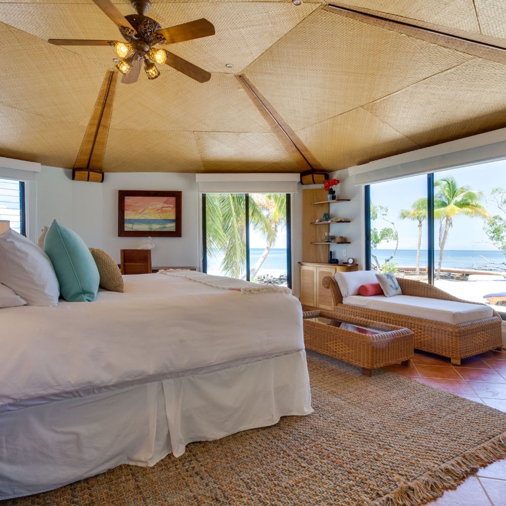 A spacious, tropical-themed bedroom featuring a large bed with white linens, decorative pillows, and a ceiling fan. Large windows provide a view of palm trees and the ocean, while a comfortable lounge chair is positioned beside the bed. Natural light fills the room, enhancing the warm color palette.