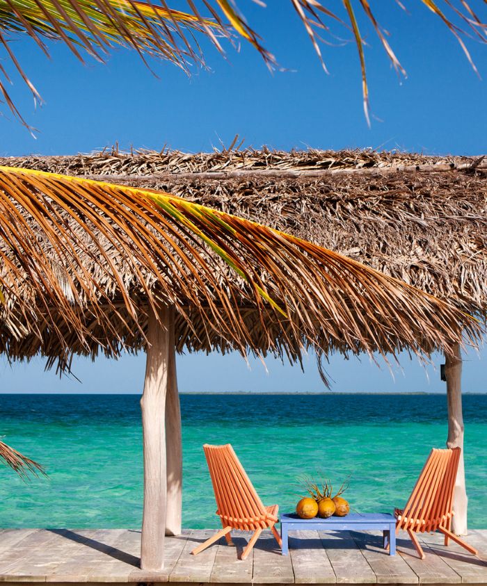 A thatched beach hut overlooking turquoise waters, with two orange lounge chairs and a small table featuring coconuts. Palm fronds frame the scene under a clear blue sky.