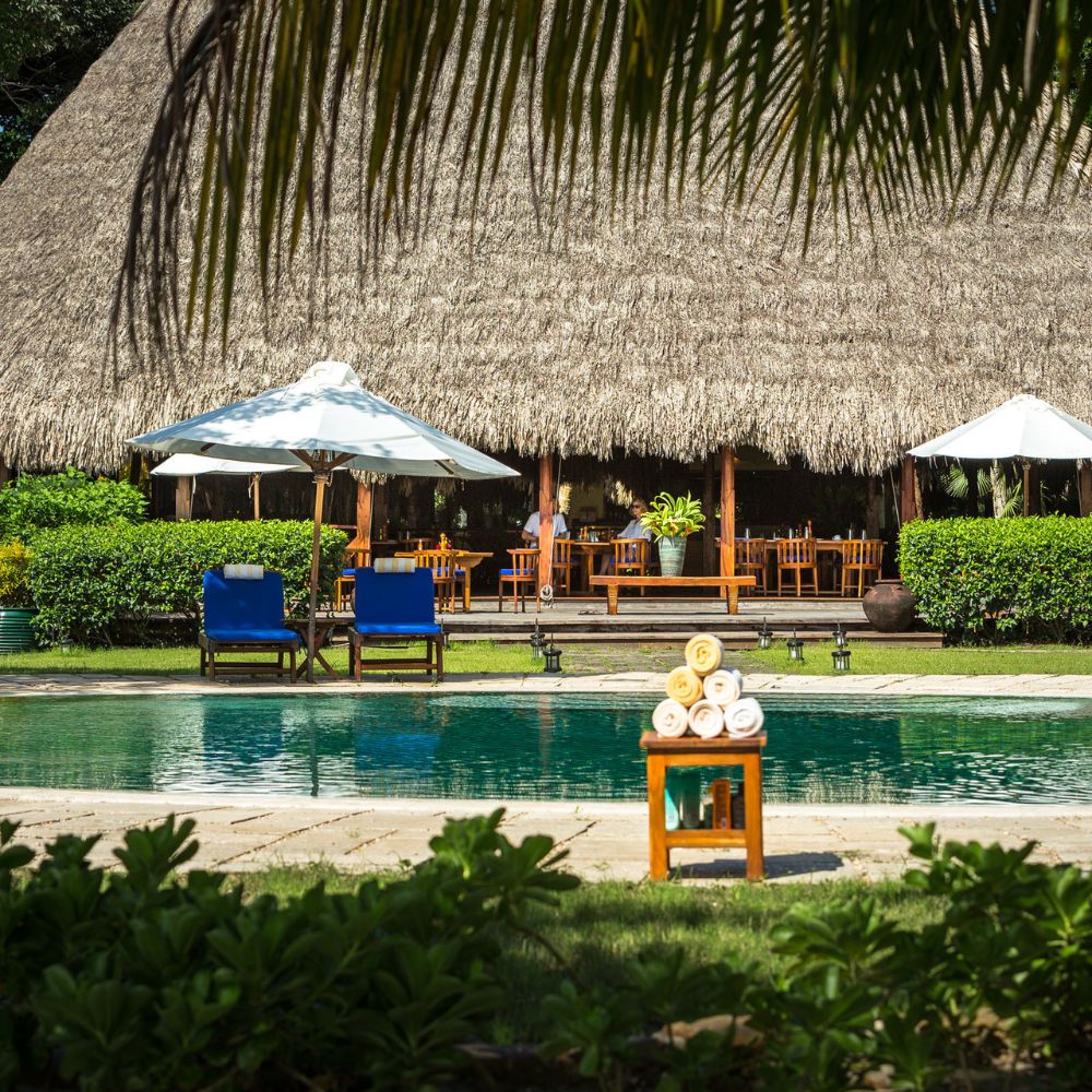 A serene outdoor scene featuring a swimming pool in the foreground, with neatly stacked towels on a small wooden table. In the background, a thatched-roof building with dining tables and umbrellas is surrounded by lush greenery.