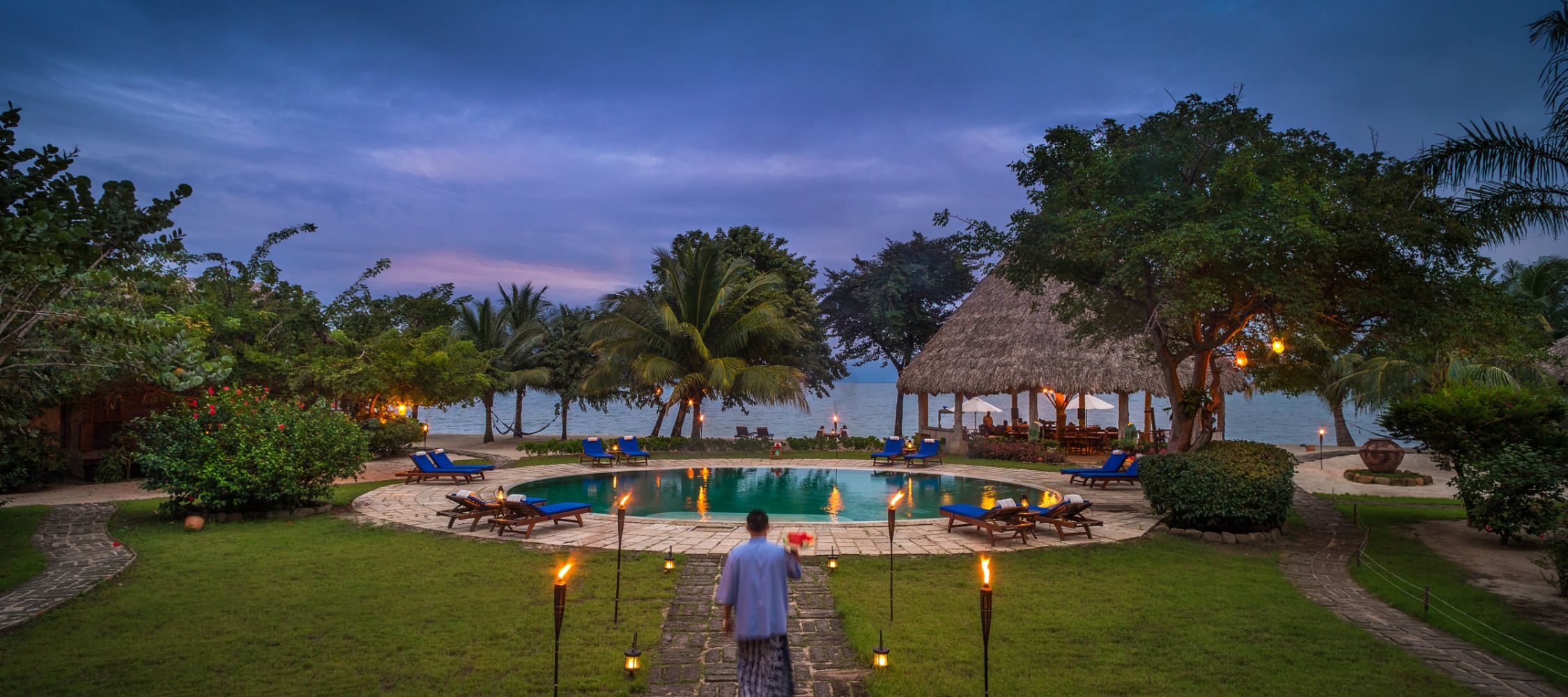 A serene tropical setting features a circular pool surrounded by green grass and palm trees. In the background, a thatched-roof hut is visible along the water's edge, with soft evening light illuminating the scene. A person walks along a stone path towards the pool, which is lined with lounge chairs and torches.