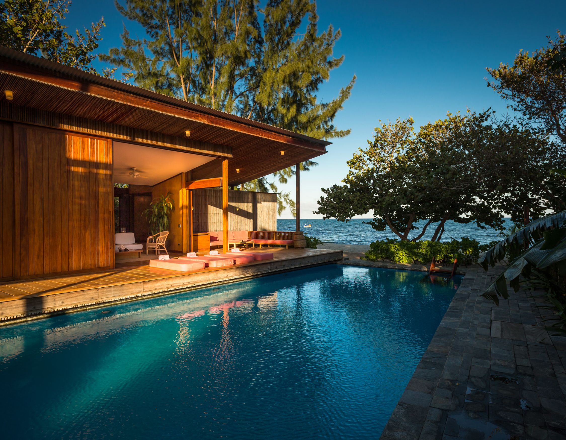 A modern wooden house with large windows overlooks a serene pool and the ocean. Lush greenery surrounds the area, creating a tranquil outdoor setting.