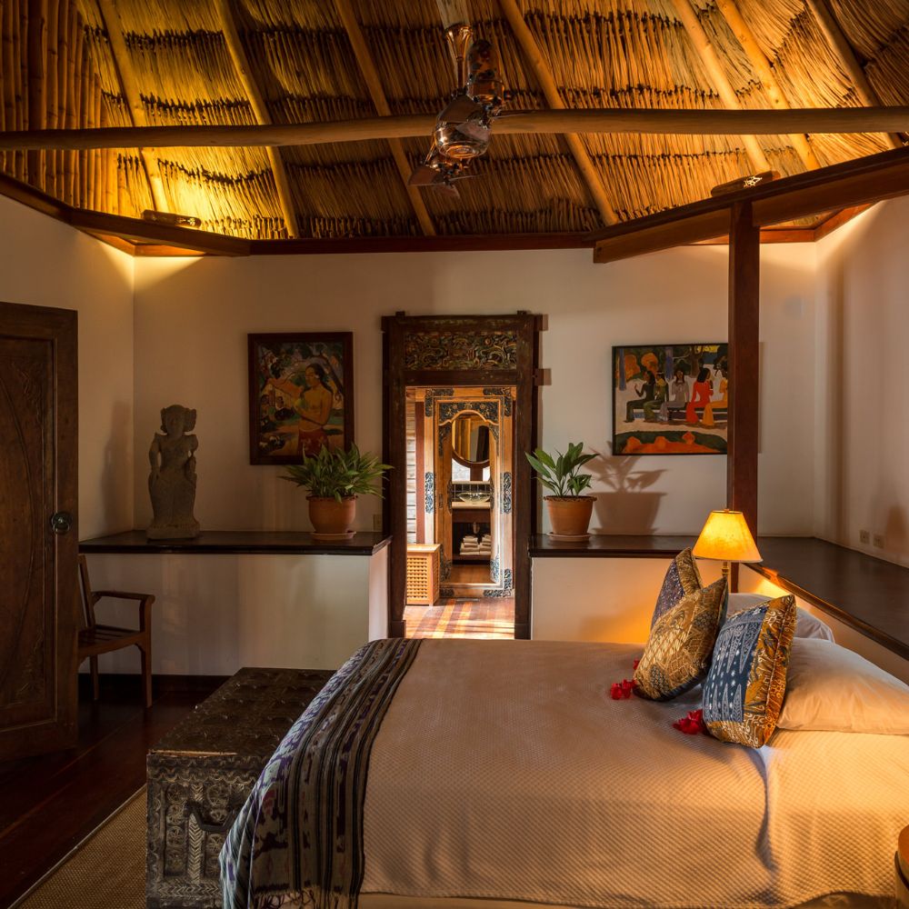 A cozy bedroom featuring a thatched ceiling, wooden furniture, and tropical decor. A comfortable bed with decorative pillows is positioned in the foreground, illuminated by a bedside lamp. Plants enhance the ambiance, while artwork adorns the walls. A doorway leads to another room in the background.