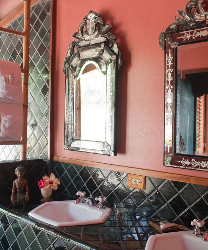 A bathroom with terracotta walls, featuring a vintage ornate mirror above a white sink. The backsplash is made of dark green tiles. A small statue and flowers are placed on the countertop.