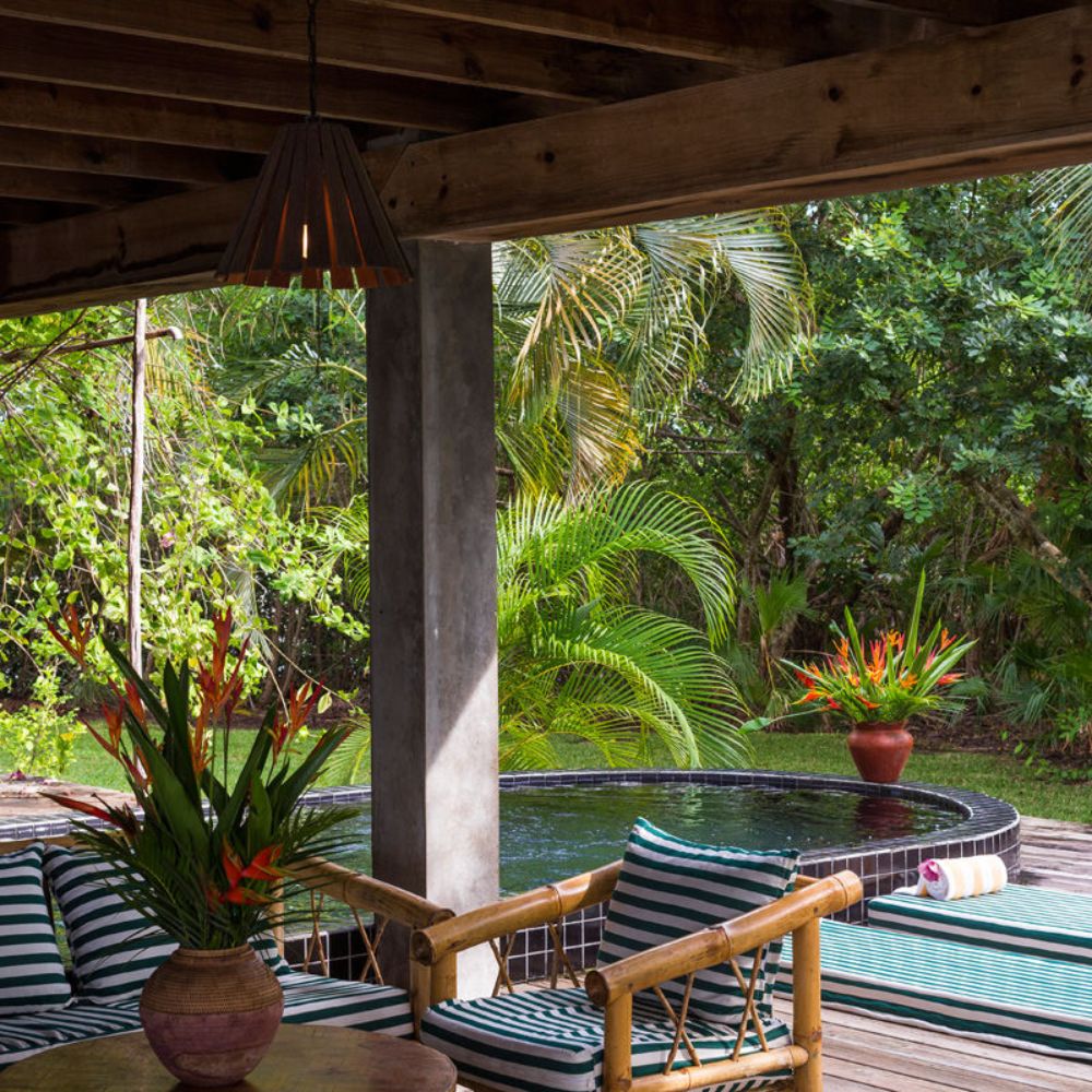 A cozy outdoor space featuring striped cushions on wooden furniture, surrounded by lush greenery. A small, dark tiled pool is visible in the background, with a potted plant adding vibrant color. Natural light filters through the structure, creating a serene atmosphere.