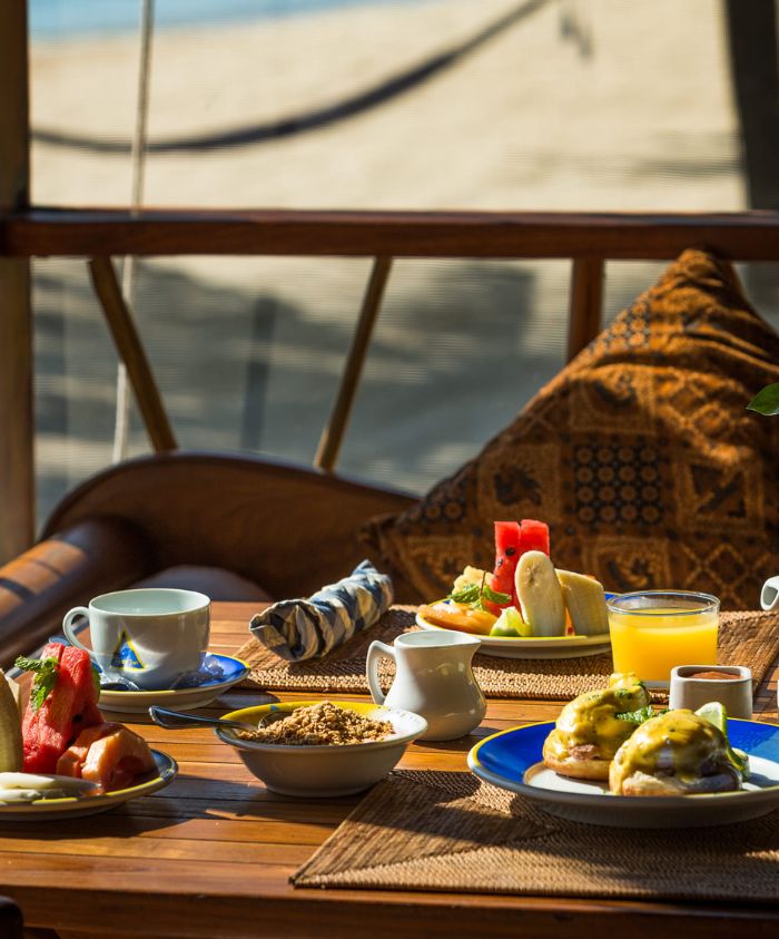 A beautifully arranged breakfast table featuring eggs benedict, fresh fruit, yogurt with granola, and beverages, set against a backdrop of a beach view.