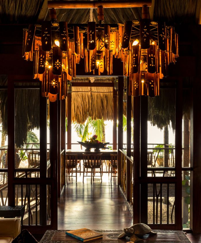 A wooden chandelier made of carved pieces hangs from a thatched ceiling, illuminating a cozy interior leading to an outdoor dining area with tropical views.