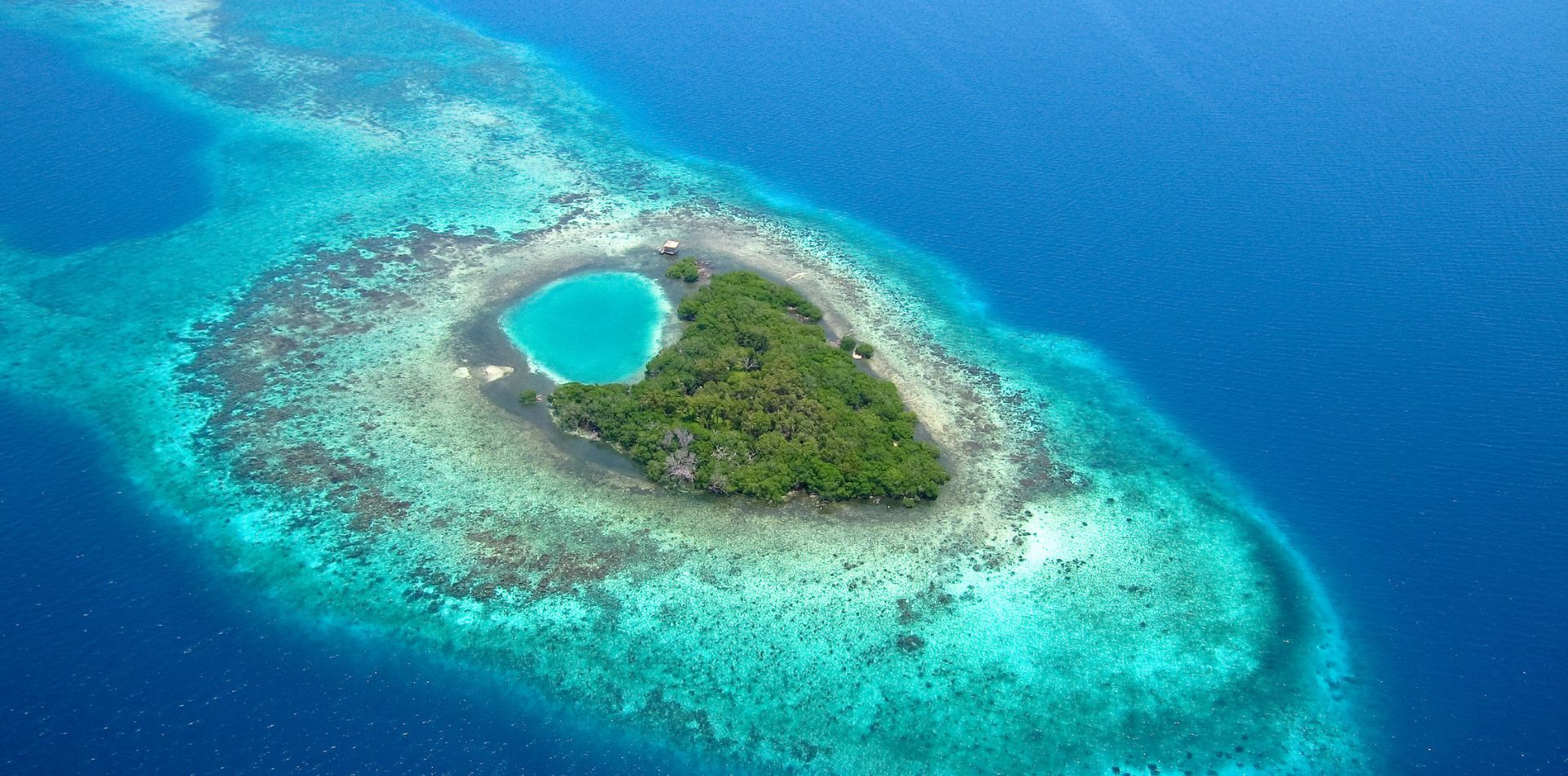 A small, lush island surrounded by vibrant turquoise waters, featuring a heart-shaped lagoon in the center and coral reefs visible beneath the surface.