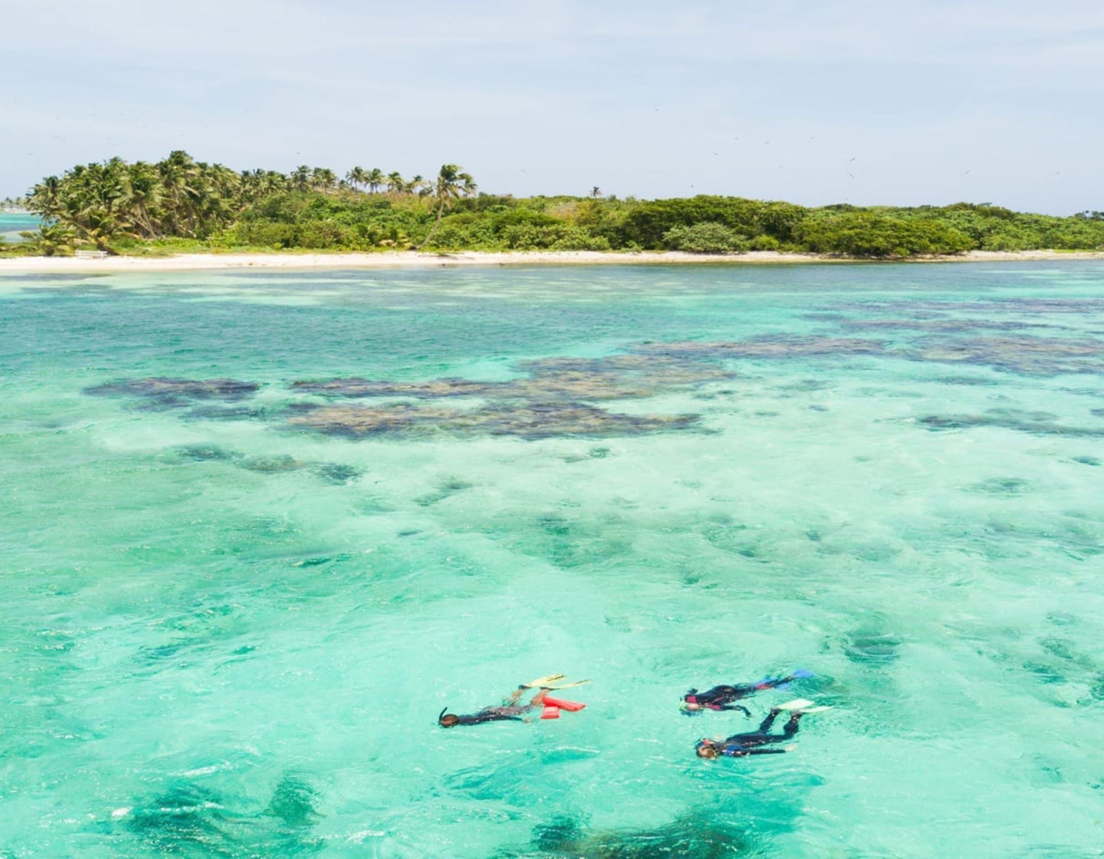 Three snorkelers explore vibrant turquoise waters above a coral reef, with lush green islands in the background and a clear blue sky overhead.