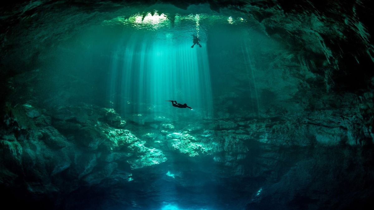 A serene underwater scene with sunlight filtering through a cave opening, illuminating the crystal-clear water. Two divers are exploring the depths, surrounded by rocky formations.