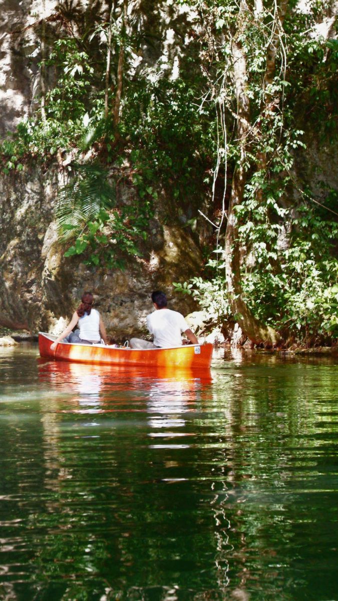 Two people paddle a vibrant orange canoe through a serene, green-tinted waterway, surrounded by lush vegetation and rocky cliffs. Sunlight filters through the trees, creating a tranquil atmosphere.