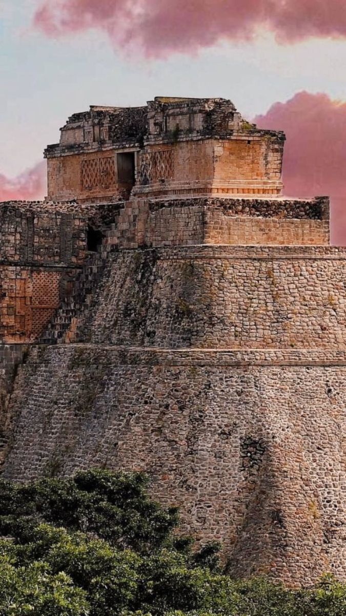 Ancient stone pyramid with a stepped design, surrounded by lush greenery and bathed in a pinkish sky.