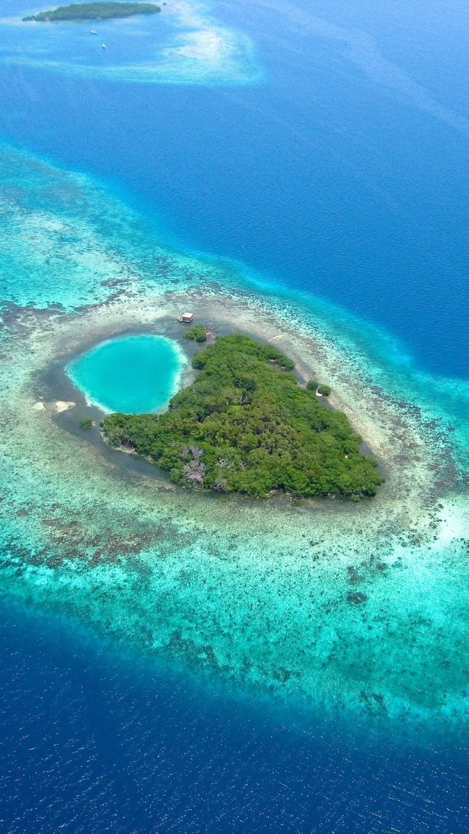 Aerial view of a small, lush tropical island surrounded by vibrant blue waters and coral reefs, featuring a circular lagoon in the centre.
