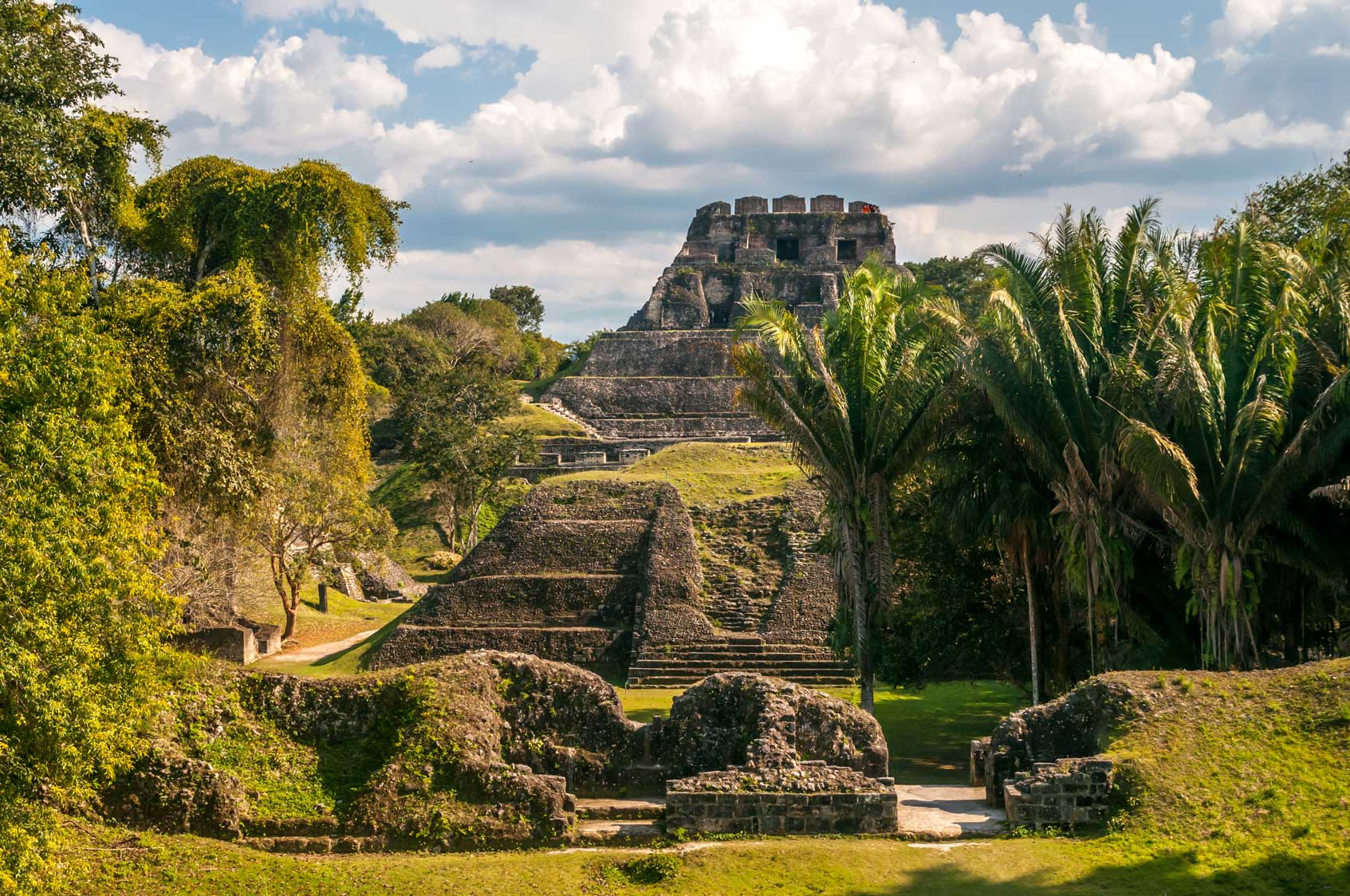 Ancient pyramid structure surrounded by lush greenery and palm trees under a partly cloudy sky.