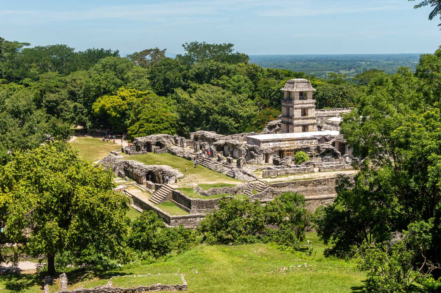 Ancient ruins surrounded by lush greenery, with stone structures and a tiered tower visible in the background. The landscape features a clear view of the horizon.