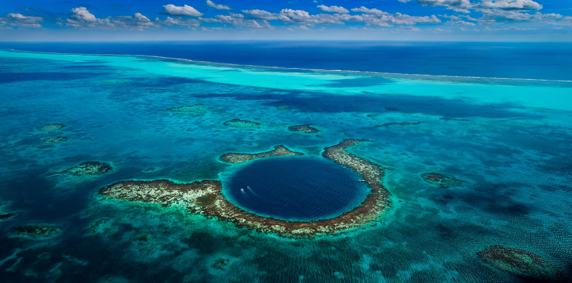 Aerial view of a circular blue hole surrounded by coral reefs in a vibrant turquoise sea under a blue sky with clouds.