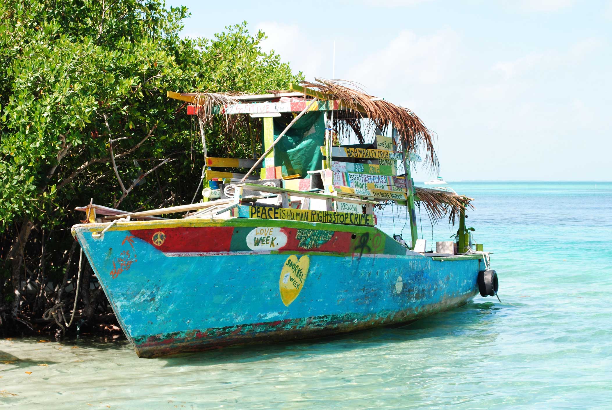 A brightly painted fishing boat with colorful signage rests by a shore, surrounded by greenery and clear blue waters. The boat features a thatched roof and various decorations, creating a vibrant, tropical atmosphere.