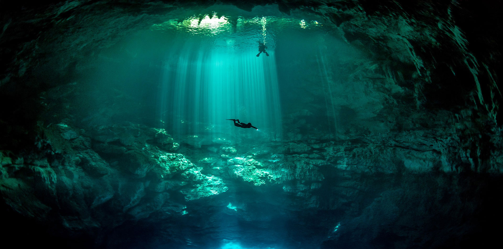 A diver swims in a deep underwater cave illuminated by beams of light piercing through the water's surface, showcasing stunning rock formations below.