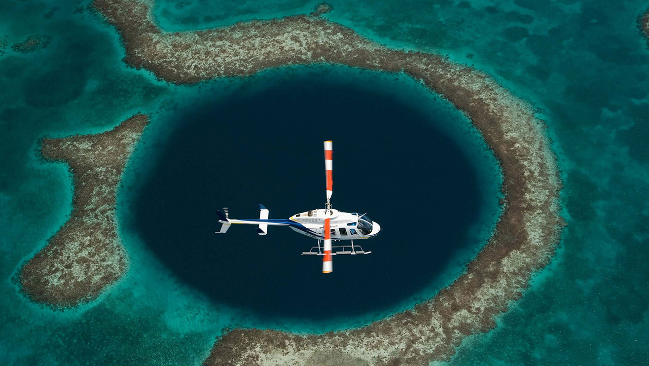 A helicopter hovers above a large circular blue hole surrounded by coral reefs, with vibrant turquoise waters.