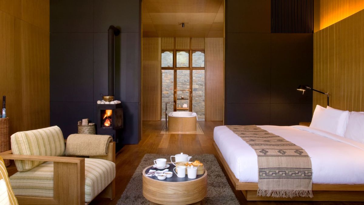 A cozy room with a large bed featuring white linens, a wooden chair with striped upholstery, and a round coffee table with snacks and tea. A small fireplace adds warmth, and large windows provide a view of a stone-walled bathroom. Warm lighting enhances the inviting atmosphere.