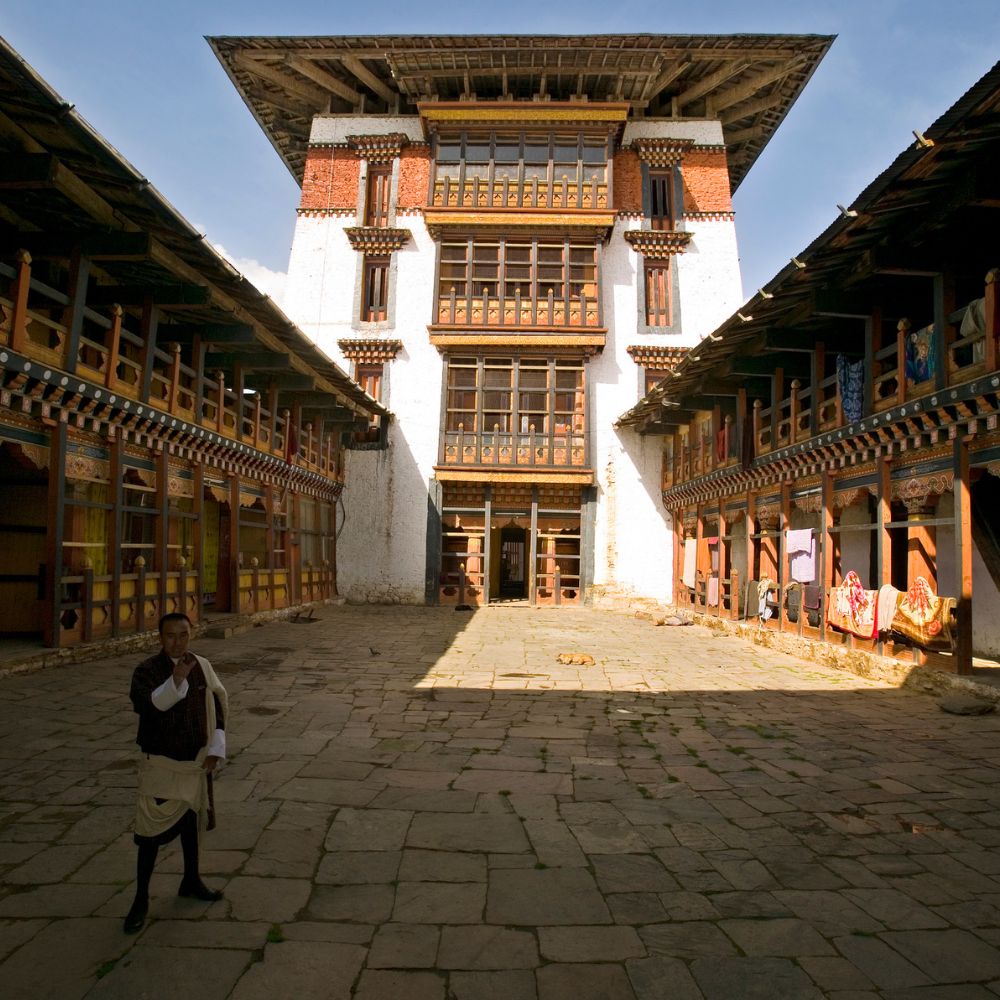 A traditional Bhutanese courtyard with a large multi-story building at the center. The building features intricate wooden carvings and brightly painted balconies. A person stands in the courtyard, surrounded by stone paving and open areas, with hanging textiles displayed nearby.