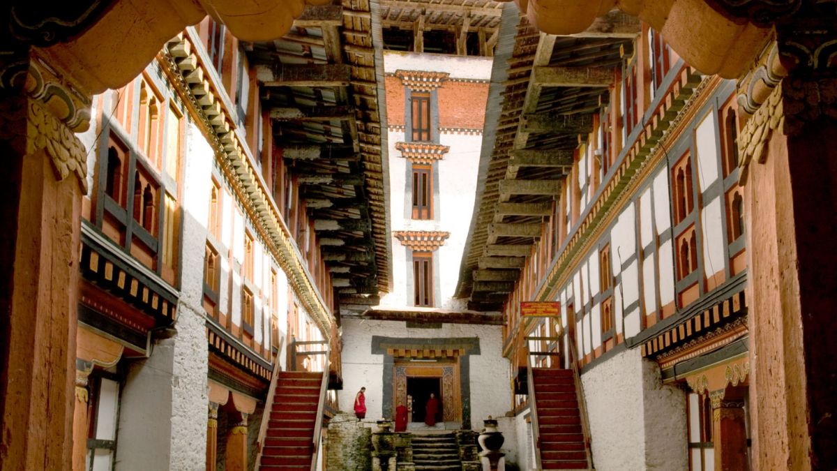 A grand courtyard of a Bhutanese monastery, featuring intricate wooden balconies and staircases. Monks in traditional robes can be seen near the entrance, surrounded by whitewashed walls and vibrant wooden accents. The atmosphere is serene and steeped in cultural heritage.