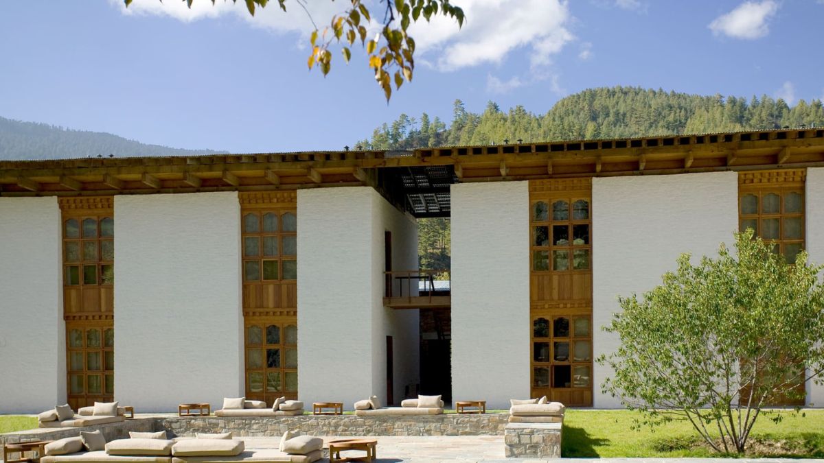 A modern building featuring a white exterior with wooden accents, large windows, and a flat roof. It is set against a backdrop of green hills and a clear blue sky. Comfortable outdoor seating is arranged in front of the structure.