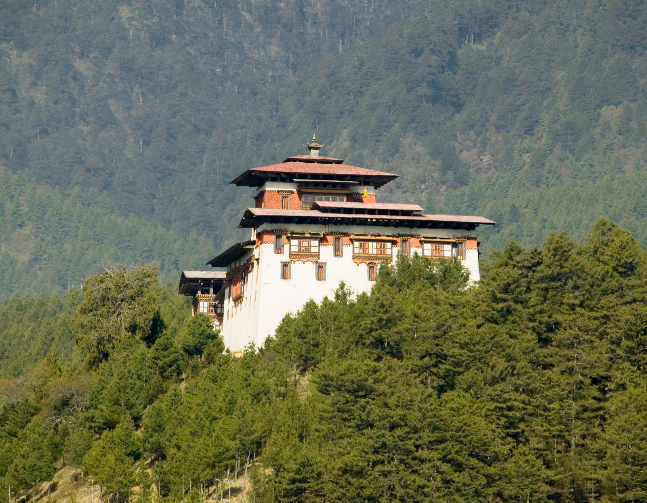 A traditional Bhutanese building situated on a hillside, surrounded by lush green trees and mountains in the background. The structure features intricate detailing and a multi-tiered roof with a decorative spire.