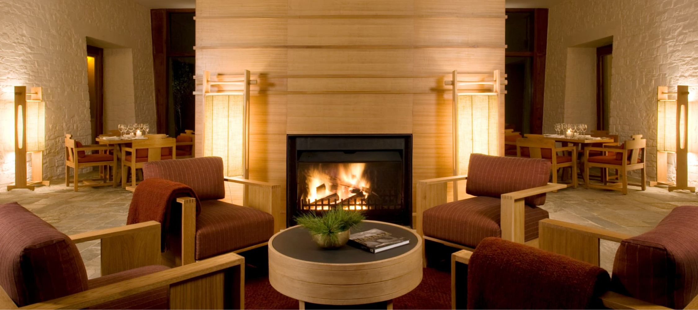 A cozy lounge area featuring two wooden armchairs with maroon cushions, a round black coffee table, and a glowing fireplace at the center. Soft lighting from nearby lamps creates a warm ambiance in a stylish interior with stone walls.