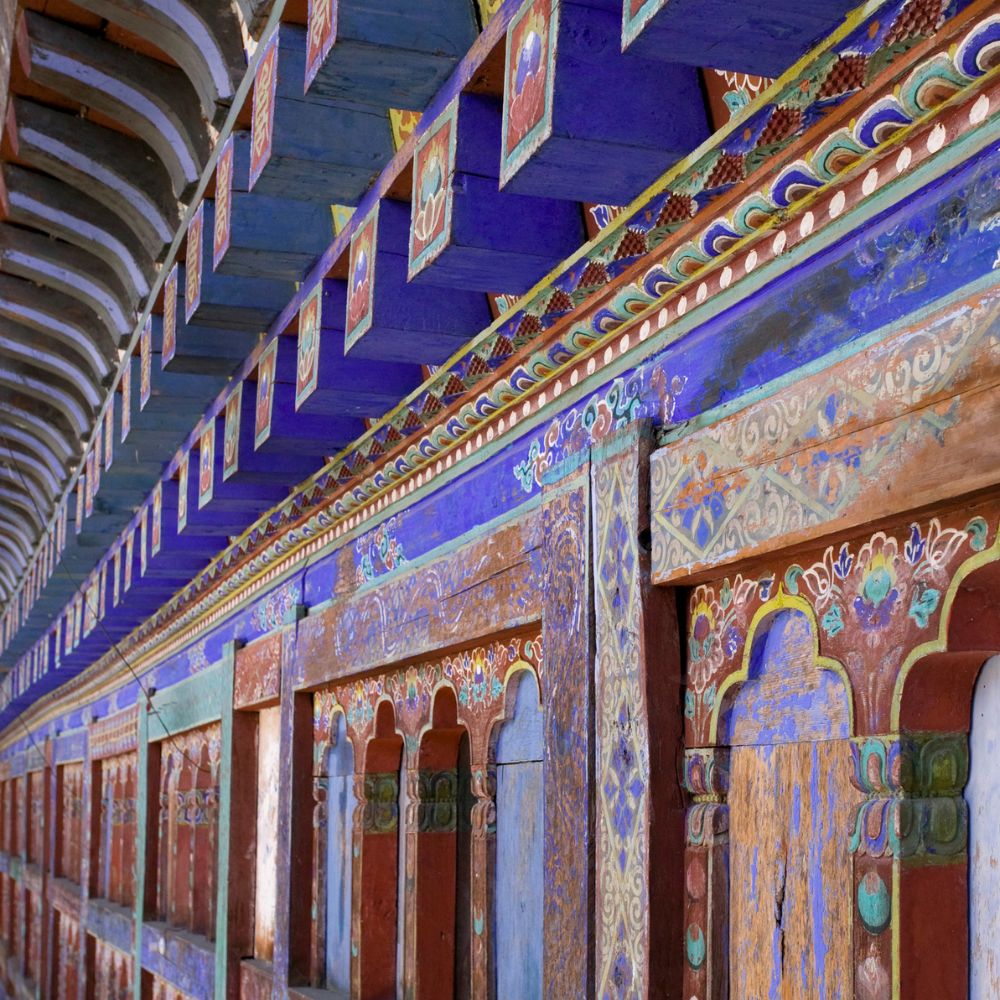 Vibrant, intricately painted wall of a temple or monastery, featuring bold blue and purple colors, ornamental designs, and detailed decorations along a corridor with arched openings.