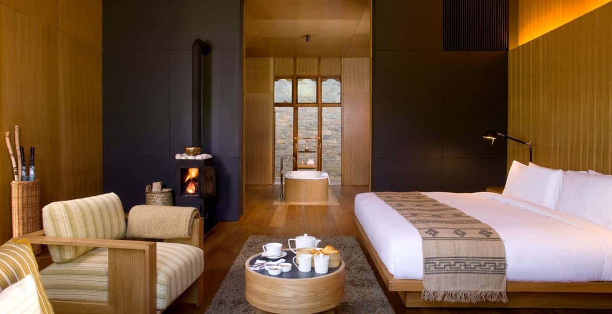 A cozy, modern hotel room featuring a large bed with white linens, a wooden floor, a comfortable seating area with striped chairs, a small round table with snacks and drinks, and a stylish bathtub in the background next to a large window. A contemporary fireplace adds warmth to the space.