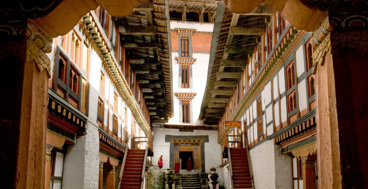 A serene courtyard of a traditional Bhutanese monastery, featuring ornate wooden architecture, sloping roofs, and staircases leading to a higher level. Red-robed monks are seen near the entrance.