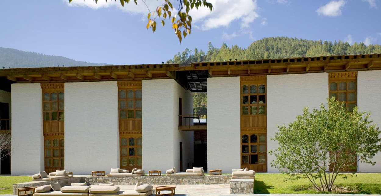 A modern building with a white facade and large wooden windows sits amid a lush green landscape, framed by mountains under a blue sky with scattered clouds. Outdoor seating is arranged on a stone patio in front of the building.