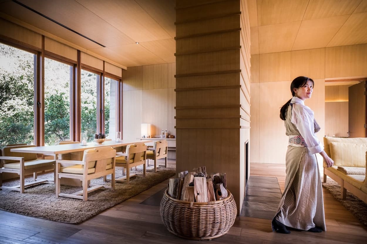 A woman in a traditional outfit walks through a modern, elegantly designed interior with wooden walls and large windows. A dining table is set nearby, and a basket of firewood sits on the floor, adding a cozy touch to the space.