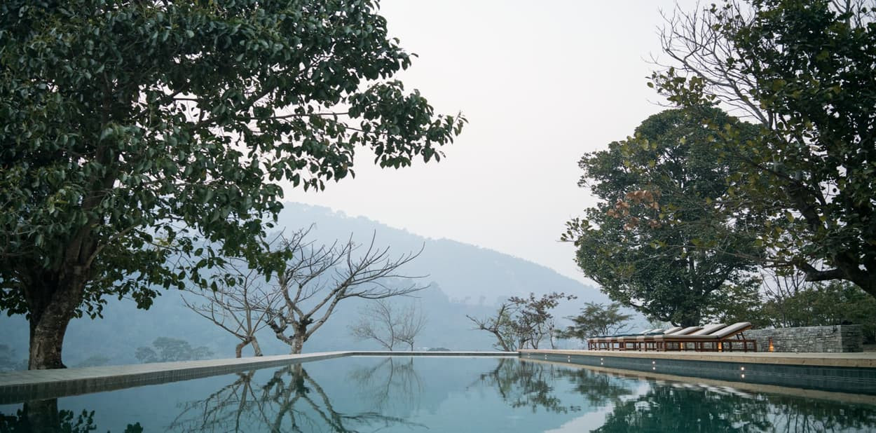 A tranquil swimming pool surrounded by lush trees and distant mountains under a hazy sky. Loungers are placed beside the pool, inviting relaxation.