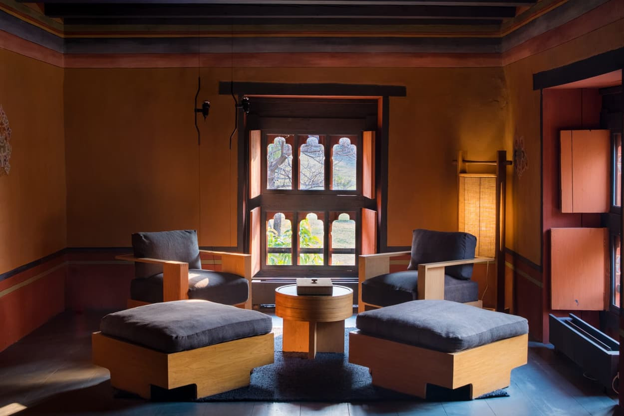A cozy room with warm orange walls, featuring two upholstered chairs and two ottomans around a round wooden table. A large window allows natural light to illuminate the space, complemented by a floor lamp.