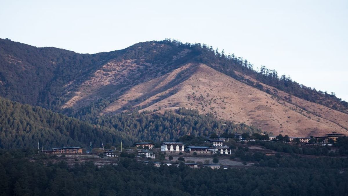 A mountainous landscape with a prominent peak in the background, featuring a cluster of traditional Bhutanese buildings nestled among lush greenery. The sky is clear, suggesting early morning or late afternoon light.