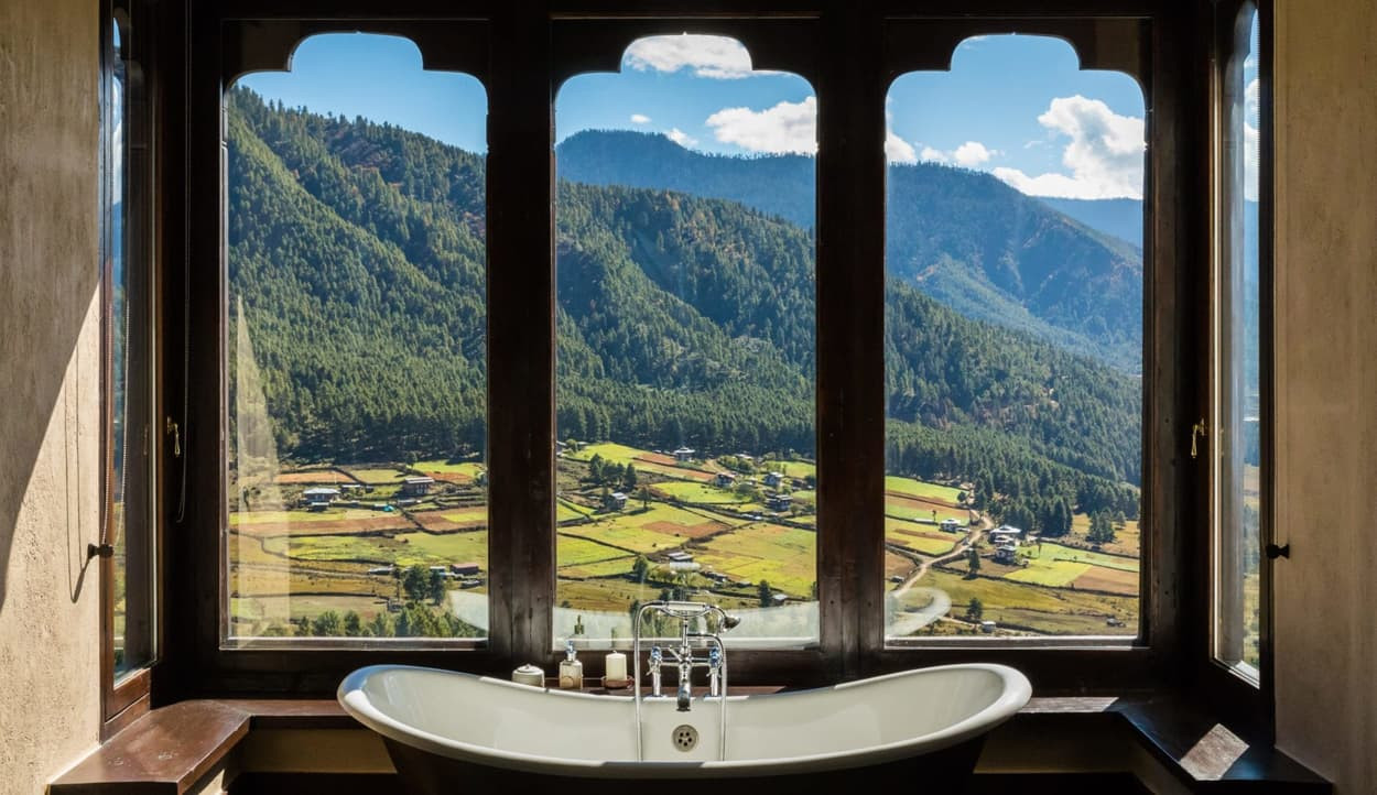 A luxurious bathtub sits near a large window, offering a stunning view of lush green mountains and terraced fields under a bright blue sky with fluffy clouds.