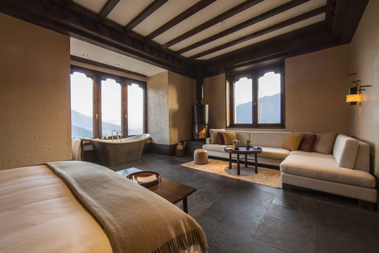A spacious hotel room with a large bed, cozy seating area, and a freestanding bathtub. Large windows offer a scenic view of mountains. A small table sits in the center, and a fireplace adds warmth to the ambiance. Earthy tones and natural materials create a serene atmosphere.