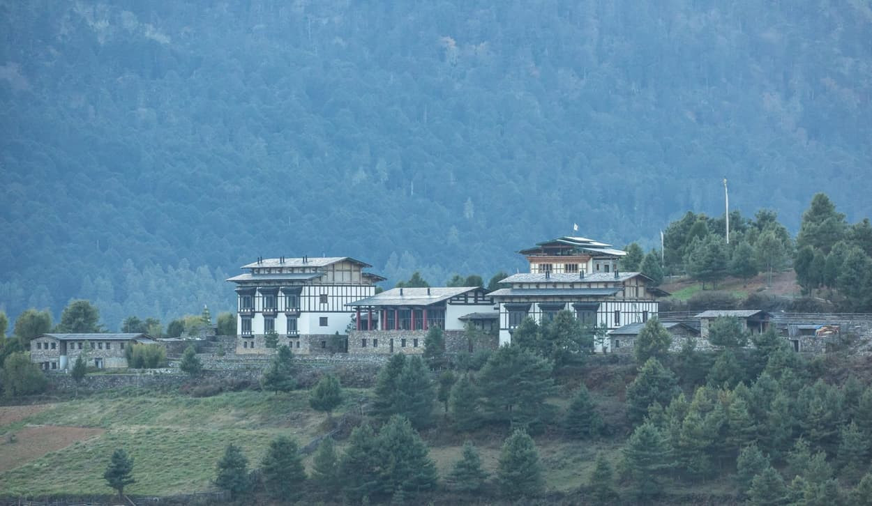 A traditional Bhutanese building set against a backdrop of mountains and pine trees, showcasing intricate architectural details and multiple stories.