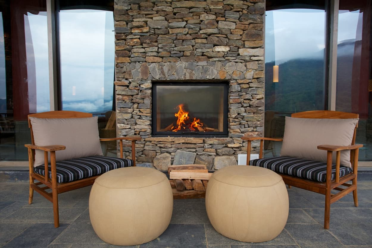 Two wooden chairs with beige cushions positioned in front of a stone fireplace, featuring a small wooden table in between. The fireplace has a warm fire burning, with a serene mountainous landscape visible through large windows.