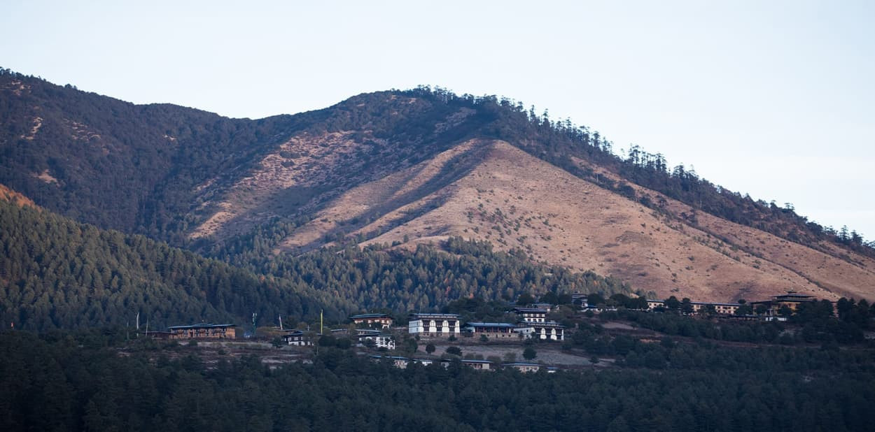 A serene mountainous landscape with a range of hills and a valley. Sparse vegetation covers the slopes, with a cluster of traditional Bhutanese buildings at the base. The scene captures the natural beauty and architecture of the region.
