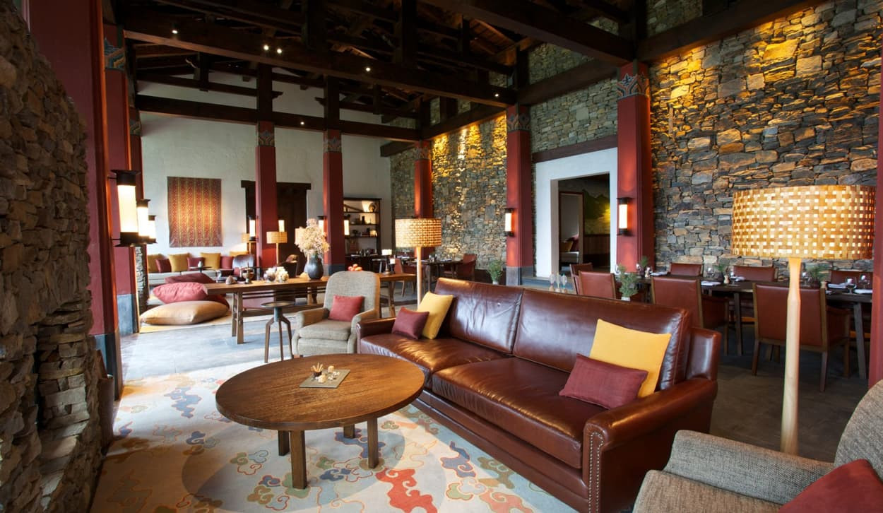 Elegant interior of a cozy lounge featuring a mix of stone walls and wooden beams. Comfortable leather sofa with colorful cushions, wooden tables, and stylish lamps create a warm atmosphere. Dining area visible in the background with rustic decor.